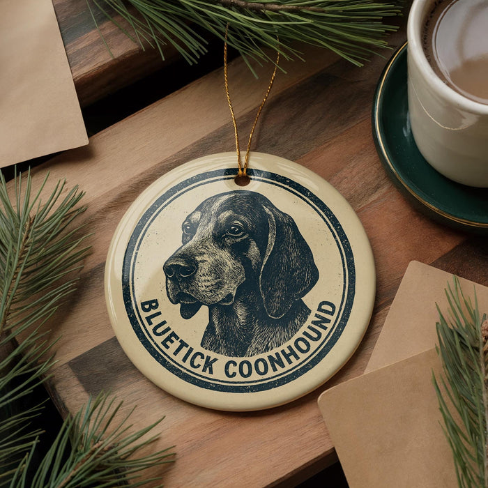 A circular ornament featuring a black and white illustration of a blue hound dog, with the text "BLUETICK COONHOUND" printed on it. The ornament is hanging from a string, and there is a cup of coffee and some pine branches visible in the background.