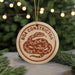 A round ornament with the text "BOA CONSTRUCTOR" and an illustration of a snake is displayed on a table, surrounded by pine branches and lights.