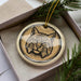A ceramic ornament in the shape of a bobcat, with a detailed illustration of the animal's face, is displayed in a box surrounded by pine branches.