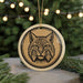A ceramic ornament featuring a black and white illustration of a bobcat's face is displayed in front of a blurred background of a Christmas tree with lights.