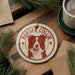 A circular ornament featuring a Border Collie dog, with the text "BORDER COLLIE" printed on it, is displayed on a wooden surface alongside a cup of coffee and some pine branches.