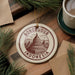 A circular ornament with the text "BOROBUDUR INDONESIA" and an illustration of Borobudur temple, which is a famous landmark in Indonesia. The ornament is hanging on a wooden surface, surrounded by pine branches and a cup of coffee.
