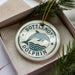 A round ornament with the text "BOTTLENOSE DOLPHIN" printed on it, surrounded by a decorative border. The ornament is placed inside a box, with pine branches visible in the background.