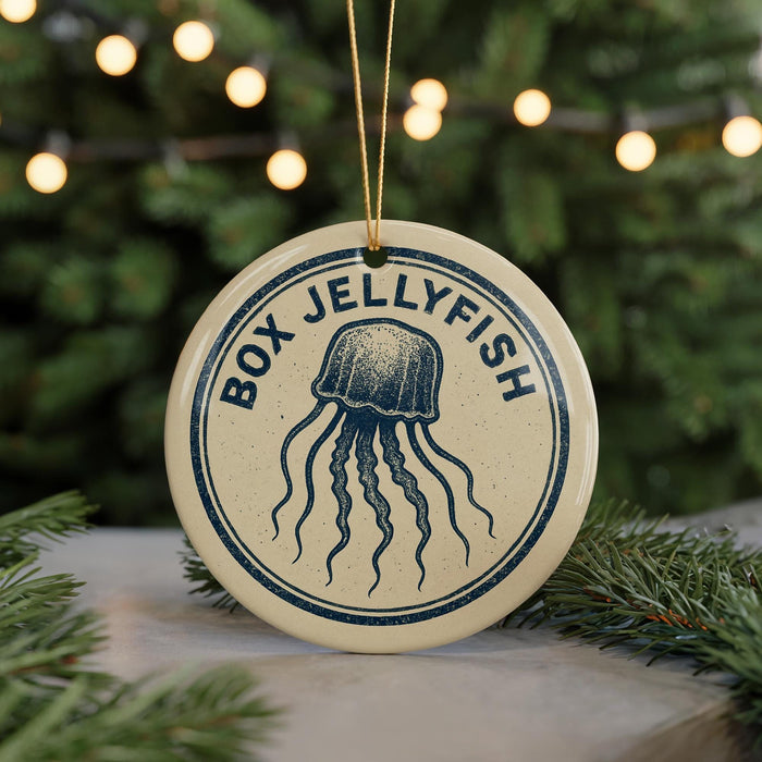 A round ornament with the text "BOX JELLYFISH" and an illustration of a jellyfish is displayed on a table, with a blurred background of a Christmas tree and lights.