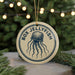 A round ornament with the text "BOX JELLYFISH" and an illustration of a jellyfish is displayed on a table, with a blurred background of a Christmas tree and lights.