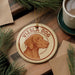 A circular ornament featuring a brown dog's head, with the text "Vizsla Dog" written around it, is displayed on a wooden surface.