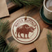 A circular ornament featuring a buffalo illustration and the text "WATER BISON" is displayed on a wooden surface, surrounded by pine branches and a cup of coffee.