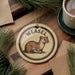 A round ornament featuring a weasel, a type of small rodent, is displayed on a wooden surface alongside a cup of coffee and some pine branches.
