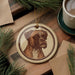 a circular ornament with a brown dog illustration on it, placed on a wooden surface next to a cup of coffee and some pine branches.