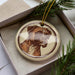 A ceramic ornament featuring a brown dog's face is displayed in a box, surrounded by pine branches.