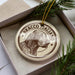 A circular ornament with a dog's head and the text "BRACCO ITALIANO" printed on it, placed inside a box with pine branches.