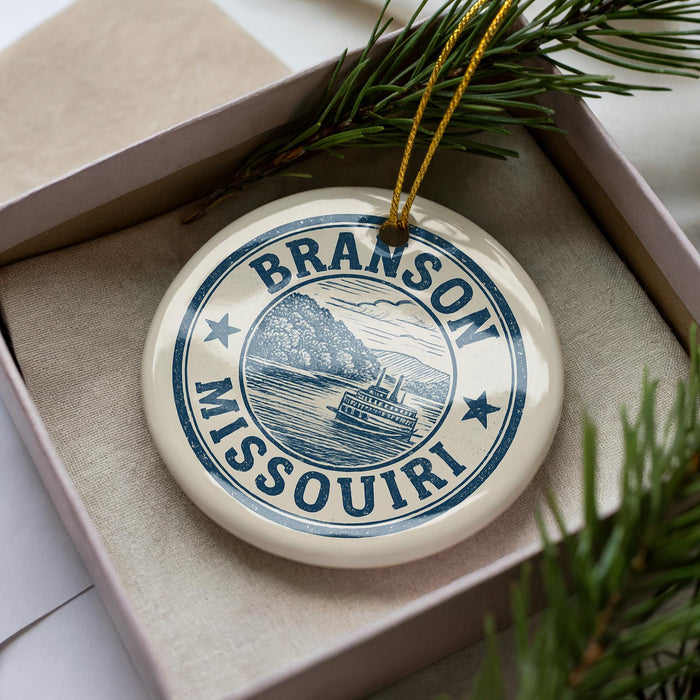 A decorative ornament in a box, featuring a blue and white design with the text "BRanson Missouri" and a depiction of a boat on a river.