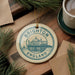 A round ornament with the text "BRIGHTON ENGLAND" and an illustration of a building, likely representing Brighton, England. The ornament is hanging from a string and is surrounded by pine branches and a cup of coffee on a wooden surface.