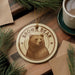 A circular ornament featuring a brown bear image is displayed on a wooden surface, surrounded by pine branches and a cup of coffee.