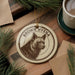 A circular ornament featuring a close-up image of a hyena's face, with the text "BROWN HYENA" printed on it, is displayed on a wooden surface alongside a cup of coffee and some pine branches.