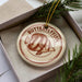 A circular ornament with a butterfly design, placed inside a box with pine branches.