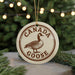 A circular ornament with the text "CANADA GOOSE" and a graphic of a goose is displayed in front of a Christmas tree with twinkling lights.