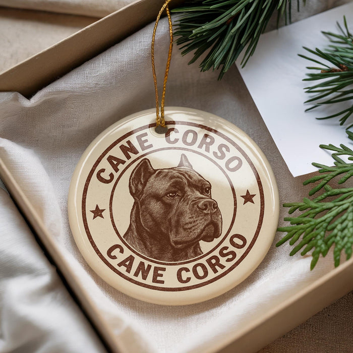 A decorative ornament featuring a brown and white image of a dog's head, with the text "CANE CORSO" printed on it. The ornament is placed inside a box with pine branches visible in the background.