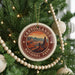 A round ornament on a Christmas tree, featuring a desert landscape with mountains and a sunset, is hanging from a string.