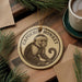 A round ornament featuring a monkey sitting on a branch, with the text "CAP PACHIN MONKEY" printed on it. The ornament is placed on a wooden surface, surrounded by pine branches and a cup of coffee.