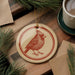 a round ornament with a red bird design, placed on a wooden surface next to a cup of coffee and some pine branches.