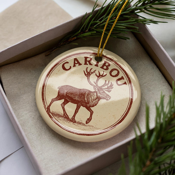 A circular ornament with a deer image and the word "CARBOU" printed on it, placed in a box with pine branches.