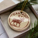 A circular ornament with a deer image and the word "CARBOU" printed on it, placed in a box with pine branches.