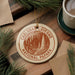 A circular ornament with the text "CARLSBAD CAVENS NATIONAL PAPARK" printed on it, surrounded by pine branches and a cup of coffee on a wooden surface.