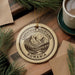 A circular ornament with the text "CARPATHIAN MOUNTAINS ROMANIA" and a mountain landscape design hangs on a wooden surface, surrounded by pine branches and a cup of coffee.