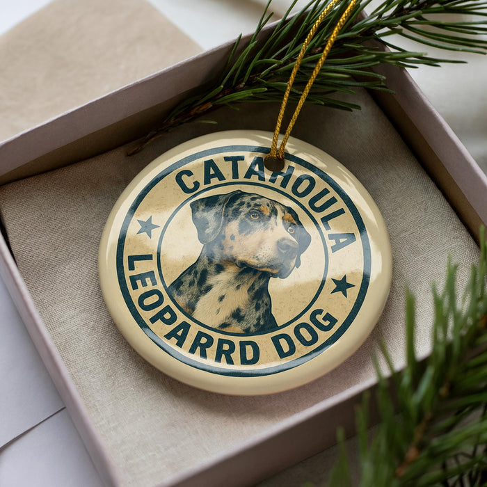 A circular ornament with a leopard dog image and the text "CATAHOULA LEOPARD DOG" printed on it, placed inside a box with pine branches.