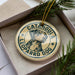 A circular ornament with a leopard dog image and the text "CATAHOULA LEOPARD DOG" printed on it, placed inside a box with pine branches.
