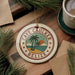 A circular ornament with the text "CAVE CAUKER BELIZE" and a palm tree design, placed on a wooden surface next to a cup of coffee and some pine branches.
