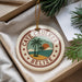 A circular ornament with the text "CAYE CAUKER BELIZE" and a palm tree design, placed on a white surface next to pine branches.