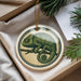 A decorative ornament featuring a chameleon is displayed in a box, surrounded by pine branches and a white card.