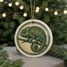 A round ornament featuring a chameleon perched on a branch, with a blurred background of pine trees and twinkling lights.