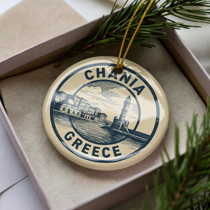 A circular ornament with the text "CHANIA GREECE" printed on it, surrounded by a decorative border. The ornament is placed inside a box, with pine branches visible in the background.