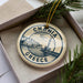 A circular ornament with the text "CHANIA GREECE" printed on it, surrounded by a decorative border. The ornament is placed inside a box, with pine branches visible in the background.