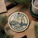 A decorative ornament featuring the text "CHANIA GREECE" and an illustration of a lighthouse, placed on a wooden surface alongside a cup of coffee and pine branches.