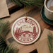 A decorative ornament featuring the city of Chengdu, China, with a mountain and pagoda in the background. The ornament is hanging from a string and is placed on a wooden surface, surrounded by pine branches and a cup of coffee.