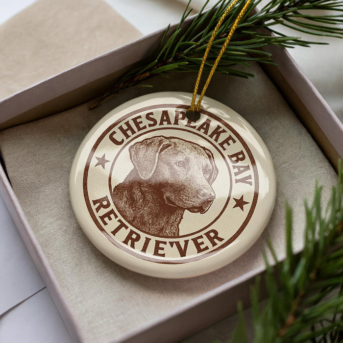 A round ornament with a brown and white design featuring a dog's head and the text "Chesapeake Bay Retriever" is displayed in a box with pine branches.