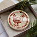 A ceramic ornament in the shape of a chicken is displayed in a box, surrounded by pine branches.