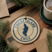 A round ornament with the text "CHILO E ISLAND CHILE" printed on it, surrounded by pine branches and a cup of coffee on a wooden surface.
