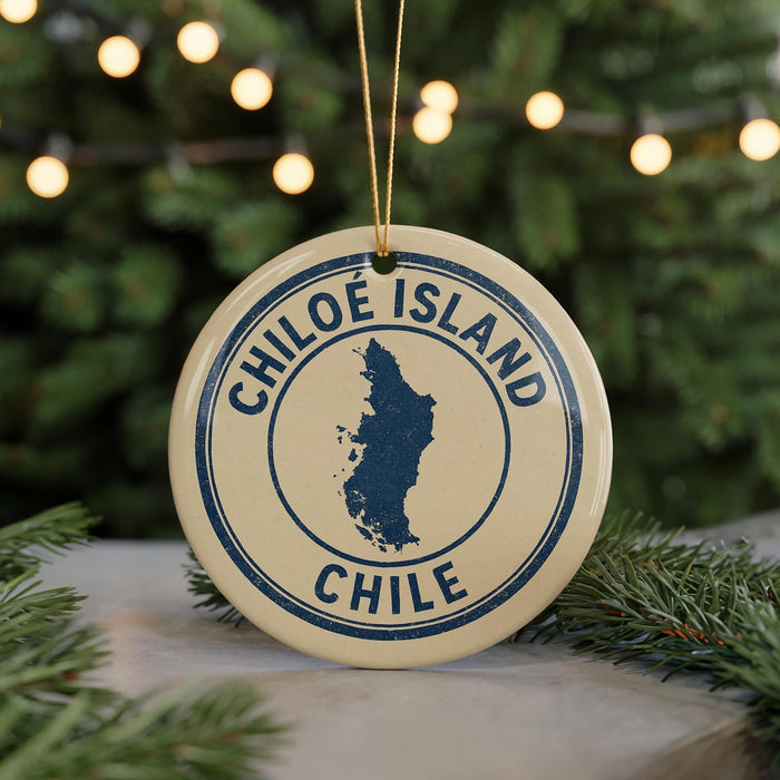 A circular ornament with the text "CHILÉ ISLAND CHILE" printed on it, surrounded by a blue border and a map of Chile. The ornament is hanging from a string, and there are pine branches visible in the background.