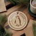A round ornament featuring a chin strap penguin is displayed on a wooden surface, surrounded by pine branches and a cup of coffee.