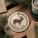 A ceramic ornament featuring a deer with antlers, surrounded by pine branches and a cup of coffee on a wooden surface.