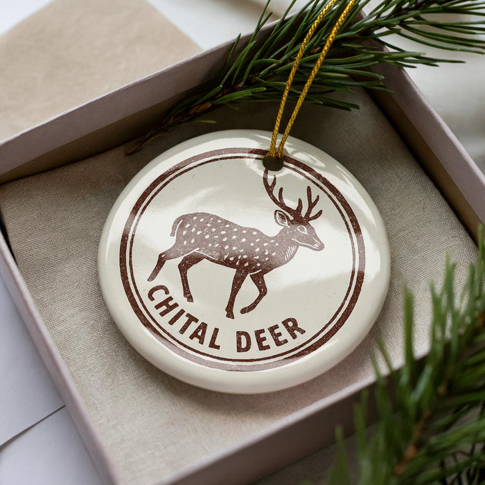 A white ceramic ornament with a brown deer illustration and the text "CHITAL DEER" is displayed in a box with pine branches.
