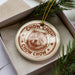 A round ornament with a chow Chow dog image and the text "CHOW CHOW" printed on it, placed inside a box with pine branches.