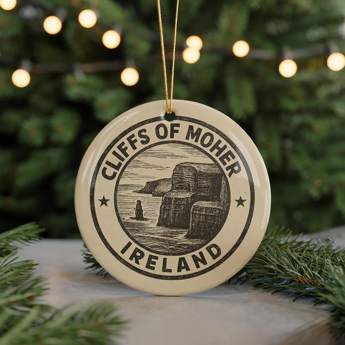 A circular ornament with the text "CLIFFS OF MOHER IRELAND" and an illustration of cliffs, hanging from a string against a background of Christmas lights and greenery.