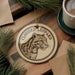 A round ornament featuring a leopard's head is displayed on a wooden surface, surrounded by pine branches and a cup of coffee.