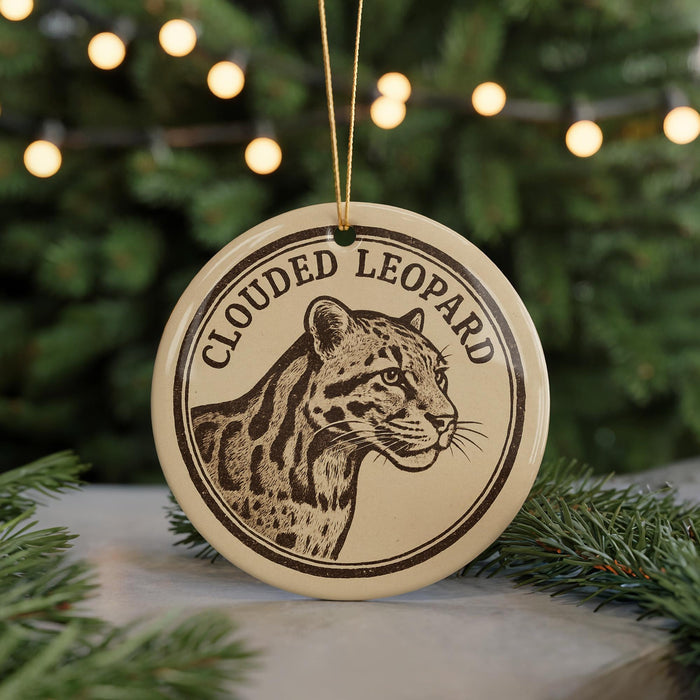 A round ornament featuring a leopard's head, with the text "CLOUDED LEOPARD" printed on it, is displayed in front of a blurred background of Christmas lights.